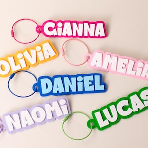 Può includere: Una collezione di sei portachiavi con nome in plastica colorata, ognuno con lettere bianche in stampatello su sfondo a tinta unita. I nomi includono Gianna, Olivia, Amelia, Daniel, Naomi e Lucas, fissati a un anello metallico circolare.