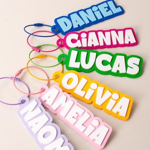 Può includere: Un set di sei etichette per bagagli rettangolari colorate con i nomi DANIEL, GIANNA, LUCAS, OLIVIA, AMELIA e NAOMI in grassetto bianco o blu. Ogni etichetta è fissata a un anello in plastica flessibile del colore corrispondente.