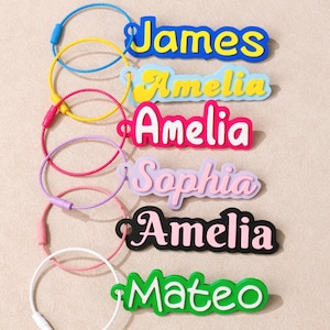 May include: A collection of six custom name keychains featuring colorful, layered acrylic nameplates attached to flexible, circular wire loops. The names James, Amelia, Sophia, and Mateo are displayed in various fonts, colors, and styles.