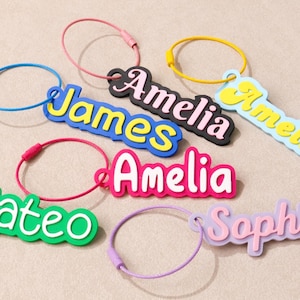 May include: A collection of colorful name keychains featuring the names James, Amelia, Mateo, and Sophia. Each keychain consists of a custom name tag attached to a circular metal wire ring in various colors including blue, pink, yellow, and purple.