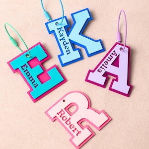 May include: Four luggage tags in the shape of the letters E, K, A, and R. Each tag features a different colour combination, with names printed on them: Emma, Kayden, Amelia, and Robert. They have matching coloured loops.