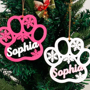 Puede incluir: Dos adornos navideños en forma de pata, uno rosa y otro blanco, cuelgan de un árbol de Navidad verde. Cada adorno presenta el nombre "Sophia" y detalles de copos de nieve. El adorno rosa tiene texto blanco, y el blanco tiene texto rosa.