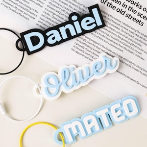 Può includere: Tre targhette personalizzate con portachiavi. Le targhette presentano i nomi "Daniel", "Oliver" e "Mateo" in lettere bianche con un contorno azzurro. Ogni targhetta ha un portachiavi di colore diverso: nero, bianco e giallo.