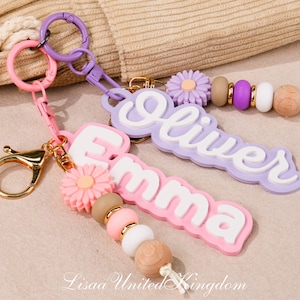 Puede incluir: Dos llaveros personalizados con placas de nombre de silicona en rosa y lavanda con letras blancas. Cada llavero incluye una cuenta en forma de flor, cuentas de silicona redondas en tonos neutros y un cierre de mosquetón de tono dorado.