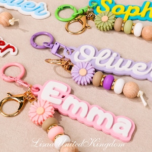 以下が含まれることがあります： 「Emma」や「Oliver」などの名前がカラフルな背景に白い文字で書かれたキーホルダー。各キーホルダーは異なる配色で、デイジーのアクセントとビーズのディテールが施されています。キーホルダーは、ゴールドまたはピンクの留め金に取り付けられています。