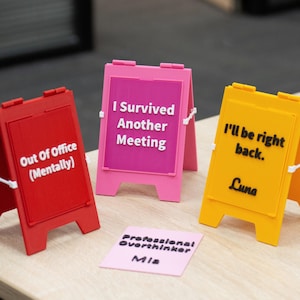 Puede incluir: Tres letreros en miniatura en forma de A en rojo, rosa y amarillo, cada uno con texto en blanco o negro: Out Of Office (Mentally), I Survived Another Meeting e I'll be right back. Luna. Un pequeño letrero rectangular rosa dice Professional Overthinker Mia.