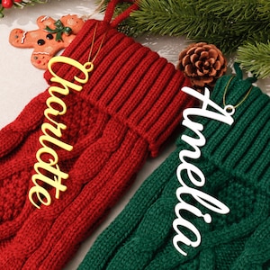 Könnte beinhalten: Ein roter gestrickter Weihnachtsstrumpf mit einem gelben Namensschild Charlotte und ein grüner gestrickter Weihnachtsstrumpf mit einem weißen Namensschild Amelia, beide mit Zopfmuster und einem umgeschlagenen Bündchen am oberen Rand.