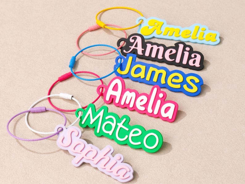 May include: A collection of six personalized name keychains featuring colorful, layered acrylic nameplates attached to thin, circular metal wire loops. The names Amelia, James, Mateo, and Sophia are displayed in various bright colors and fonts.