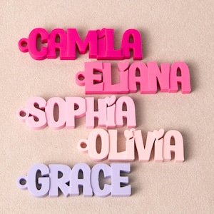 May include: Five custom name tags in varying shades of pink and purple. Each tag features a name in a bold, rounded font with a small circular hole on the left side. The names displayed are CAMILA, ELIANA, SOPHIA, OLIVIA, and GRACE in descending order.