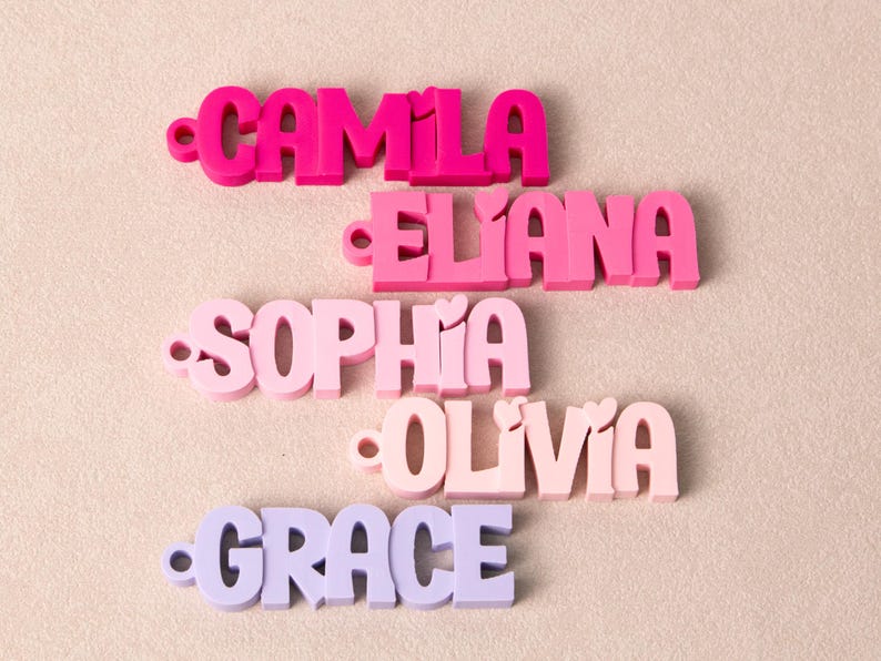 May include: Five custom name tags in varying shades of pink and purple. Each tag features a name in a bold, rounded font with a small circular hole on the left side. The names displayed are CAMILA, ELIANA, SOPHIA, OLIVIA, and GRACE in descending order.