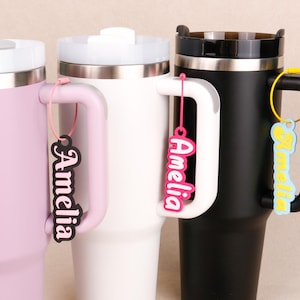 May include: Three insulated tumblers in light pink, white, and black, each featuring a handle and a lid. Attached to each handle is a personalized name tag with the name Amelia in different colors, including black, hot pink, and light blue.