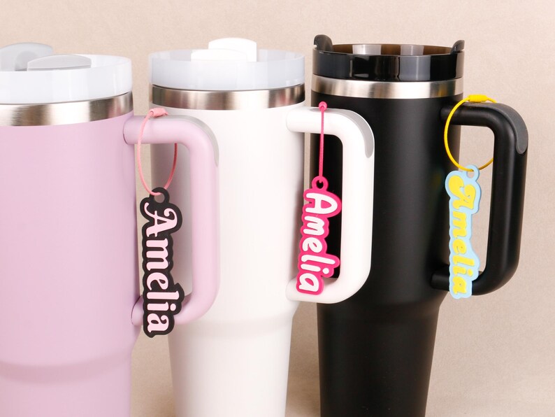 May include: Three insulated tumblers in light pink, white, and black, each featuring a handle and a lid. Attached to each handle is a personalized name tag with the name Amelia in different colors, including black, hot pink, and light blue.