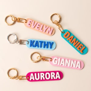 Könnte beinhalten: Fünf rechteckige Schlüsselanhänger mit Metallringen und Verschlüssen. Die Anhänger haben erhabene Schriftzüge in verschiedenen Farben: rosa EVELYN auf pfirsichfarben, orange DANIEL auf türkis, blau KATHY auf hellblau, weiß GIANNA auf hellrosa und weiß AURORA auf pink.