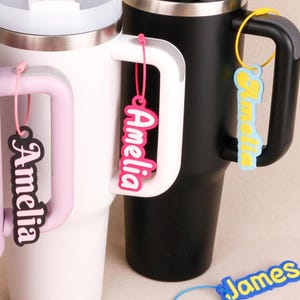 May include: Three insulated tumblers in light purple, white, and black, each featuring a handle. Attached to the handles are custom name tags with the name Amelia in various color combinations, plus a blue and yellow James name tag below.