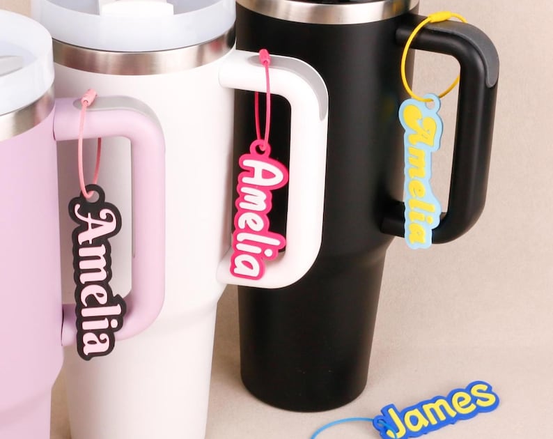 May include: Three insulated tumblers in light purple, white, and black, each featuring a handle. Attached to the handles are custom name tags with the name Amelia in various color combinations, plus a blue and yellow James name tag below.