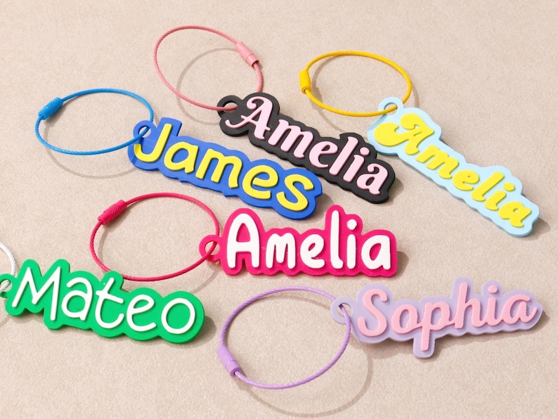 May include: A collection of colorful name keychains featuring the names James, Amelia, Mateo, and Sophia. Each keychain consists of a custom name tag attached to a circular metal wire ring in various colors including blue, pink, yellow, and purple.