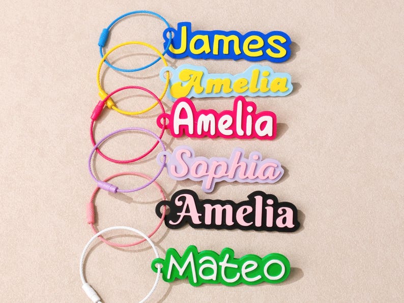 May include: A collection of six custom name keychains featuring colorful, layered acrylic nameplates attached to flexible, circular wire loops. The names James, Amelia, Sophia, and Mateo are displayed in various fonts, colors, and styles.