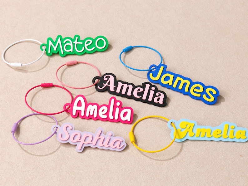 May include: A collection of six personalized luggage tags featuring names Mateo, James, Amelia, and Sophia in various colors including green, blue, black, pink, purple, and light blue, each attached to a circular wire cable loop.