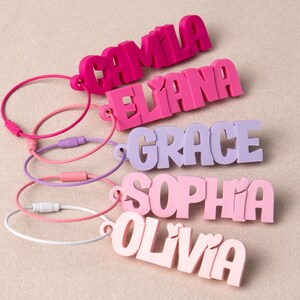 May include: Five custom name keychains in various shades of pink and purple, each featuring a 3D block letter name including CAMILA, ELIANA, GRACE, SOPHIA, and OLIVIA, attached to a circular metal wire loop with a screw-top closure mechanism.