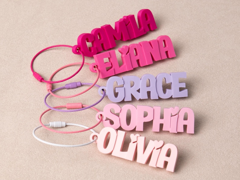 May include: Five custom name keychains in various shades of pink and purple, each featuring a 3D block letter name including CAMILA, ELIANA, GRACE, SOPHIA, and OLIVIA, attached to a circular metal wire loop with a screw-top closure mechanism.