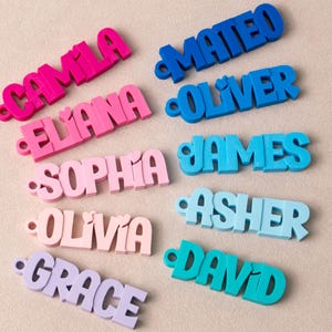 May include: A collection of ten 3D-printed name keychains in various colors. The left column features pink and purple shades with the names Camila, Eliana, Sophia, Olivia, and Grace. The right column features blue and teal shades with the names Mateo, Oliver, James, Asher, and David. Each keychain has a small circular hole on the left side.