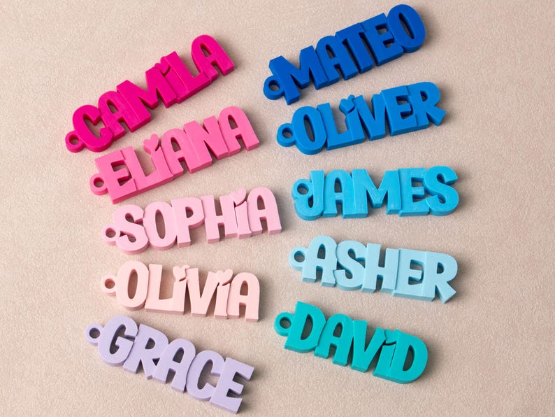 May include: A collection of ten 3D-printed name keychains in various colors. The left column features pink and purple shades with the names Camila, Eliana, Sophia, Olivia, and Grace. The right column features blue and teal shades with the names Mateo, Oliver, James, Asher, and David. Each keychain has a small circular hole on the left side.