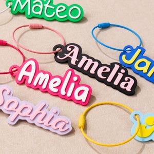 May include: A collection of custom name keychains featuring colorful, layered acrylic or silicone lettering attached to circular metal wire loops. The names Mateo, Amelia, Sophia, and James are displayed in various color combinations like green, pink, and blue.