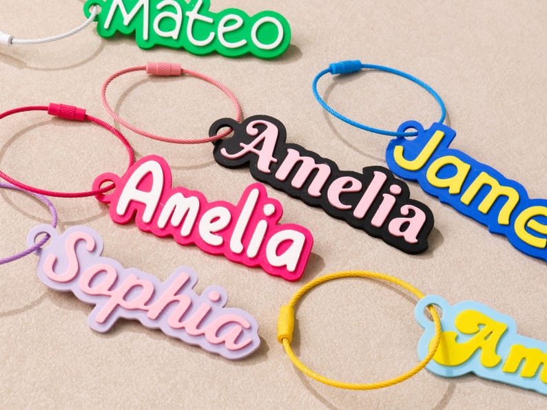 May include: A collection of custom name keychains featuring colorful, layered acrylic or silicone lettering attached to circular metal wire loops. The names Mateo, Amelia, Sophia, and James are displayed in various color combinations like green, pink, and blue.