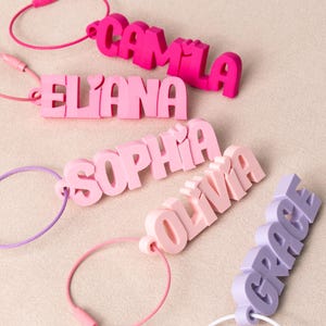 May include: Five custom name keychains in various shades of pink and purple. Each keychain features 3D block lettering with a small heart detail above the letters. The names displayed are Camila, Eliana, Sophia, Olivia, and Grace, each attached to a thin, circular metal wire loop for secure fastening.