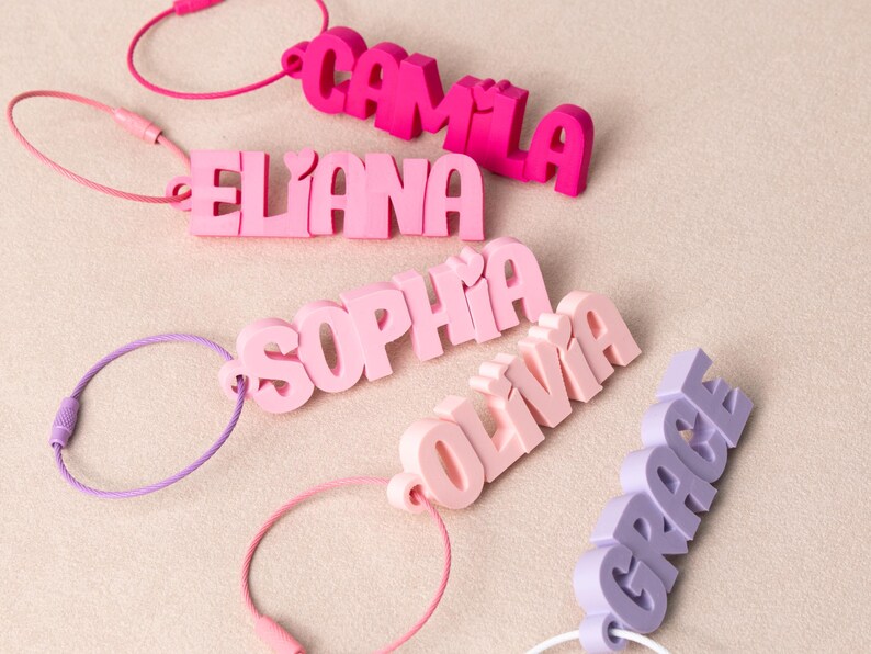 May include: Five custom name keychains in various shades of pink and purple. Each keychain features 3D block lettering with a small heart detail above the letters. The names displayed are Camila, Eliana, Sophia, Olivia, and Grace, each attached to a thin, circular metal wire loop for secure fastening.