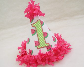 May include: A white party hat with a watermelon print and a green number one on the front. The hat is decorated with pink tissue paper.