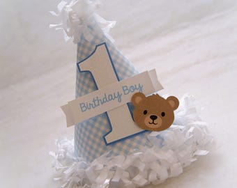 May include: Blue and white gingham party hat with a white number one and a brown teddy bear face. The hat has a white banner that says "Birthday Boy".