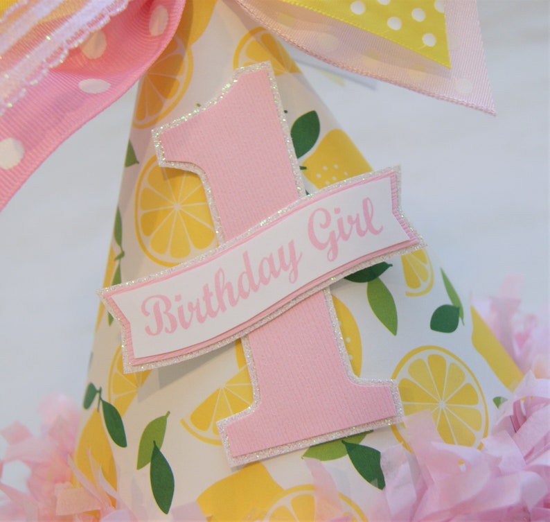May include: A pink and white party hat with a lemon print. The hat has a pink number one and a banner that says "Birthday Girl".