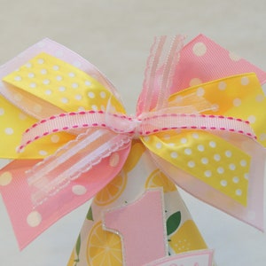 May include: A pink and yellow party hat with a large bow on top. The hat is decorated with lemons and the number one.