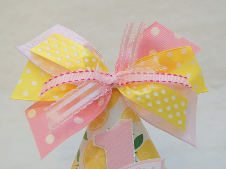 May include: A pink and yellow party hat with a large bow on top. The hat is decorated with lemons and the number one.