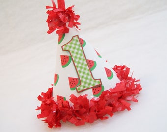 May include: A white party hat with a red fringe and a watermelon pattern. The hat has a large green and white checkered number "1" on the front.