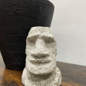 Puede incluir: Pequeña estatua Moai gris claro y desgastada, con nariz y labios prominentes. Frente a un jarrón negro grande y texturizado. Sobre una superficie de madera.