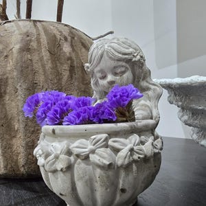 May include: A small, weathered, concrete planter shaped like a cherub holding a pot filled with vibrant purple flowers. The planter has a detailed floral design around the rim. A larger, textured pot is in the background.