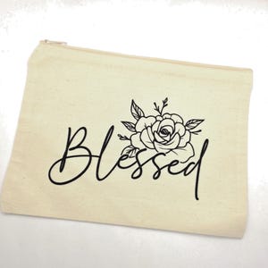 May include: A rectangular beige canvas zipper pouch featuring the word Blessed in a black cursive script font. Above the word, there is a black line art illustration of a blooming rose with leaves and stems centered on the front of the pouch.