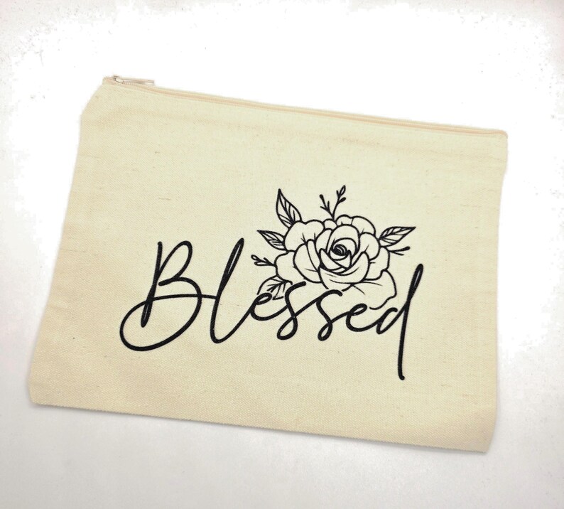 May include: A rectangular beige canvas zipper pouch featuring the word Blessed in a black cursive script font. Above the word, there is a black line art illustration of a blooming rose with leaves and stems centered on the front of the pouch.