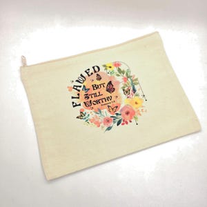 May include: A beige canvas zipper pouch featuring a floral and butterfly graphic with the text "FLAWED BUT STILL WORTHY Romans 5:8" printed in black and orange. The design includes colorful flowers and butterflies surrounding the central text.