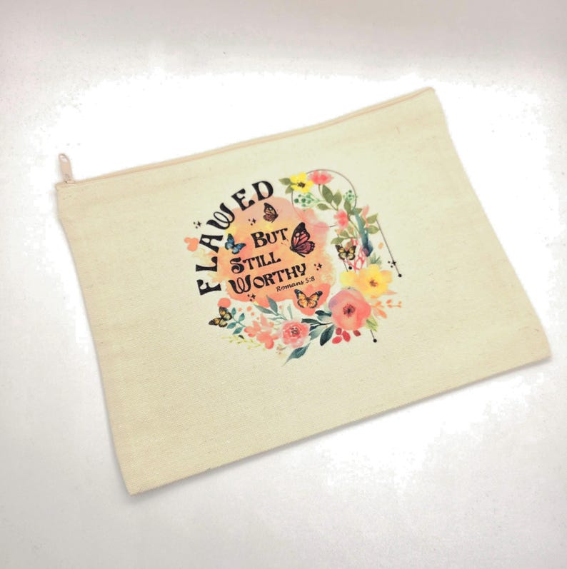 May include: A beige canvas zipper pouch featuring a floral and butterfly graphic with the text "FLAWED BUT STILL WORTHY Romans 5:8" printed in black and orange. The design includes colorful flowers and butterflies surrounding the central text.