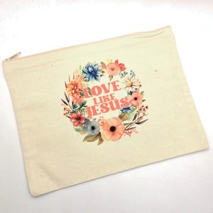 May include: A rectangular beige canvas zipper pouch featuring a floral wreath graphic in shades of pink, orange, blue, and green. Centered within the wreath is the text LOVE LIKE JESUS printed in a bold, coral-colored sans-serif font.