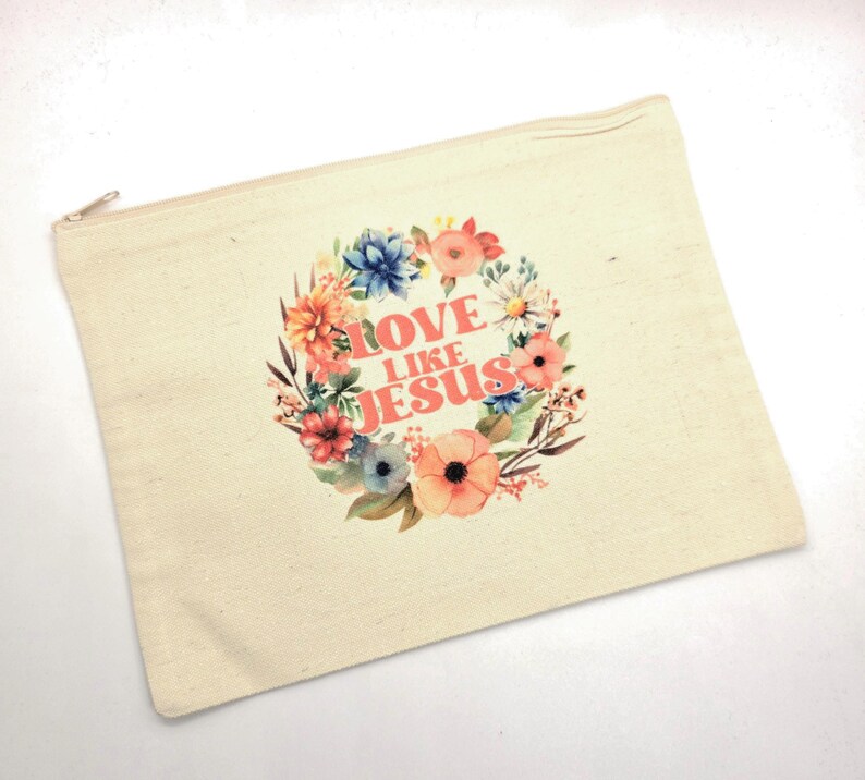 May include: A rectangular beige canvas zipper pouch featuring a floral wreath graphic in shades of pink, orange, blue, and green. Centered within the wreath is the text LOVE LIKE JESUS printed in a bold, coral-colored sans-serif font.