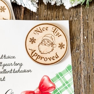 Peut inclure: Ornement de Noël en bois avec l'inscription "Nice List Approved" entourant une illustration du Père Noël. L'ornement est rond avec des détails de flocons de neige. L'image montre également un nœud rouge et un motif à carreaux verts.