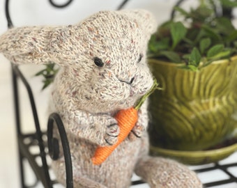 May include: A knitted gray bunny rabbit holding a small orange carrot. The bunny is sitting on a black metal shelf with green plants in a green pot behind it.