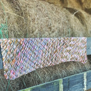 Pode incluir: Uma echarpe de malha colorida com um padrão texturizado pendurada em uma cerca de madeira com um fundo de fardos de feno.