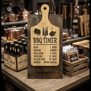 May include: A light-colored wooden cutting board shaped like a paddle, featuring a dark graphic of ribs, bottles, and a chicken, with the text BBQ TIMER and a list of cooking times measured in beers, mounted on a rectangular, weathered metal base.