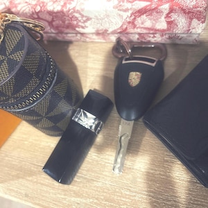 May include: An assortment of luxury items, including a cylindrical case with a gold zipper and chain, a black key fob with a Porsche emblem, a black wallet, and a black lipstick case. The items are arranged on a light wood surface.