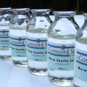 May include: A row of six clear glass bottles with black lids and blue and white labels. The labels read "Concord Sc" and "Warm Vanilla Scent" with the text "Fragrance Oil" and "Weight 1 oz".